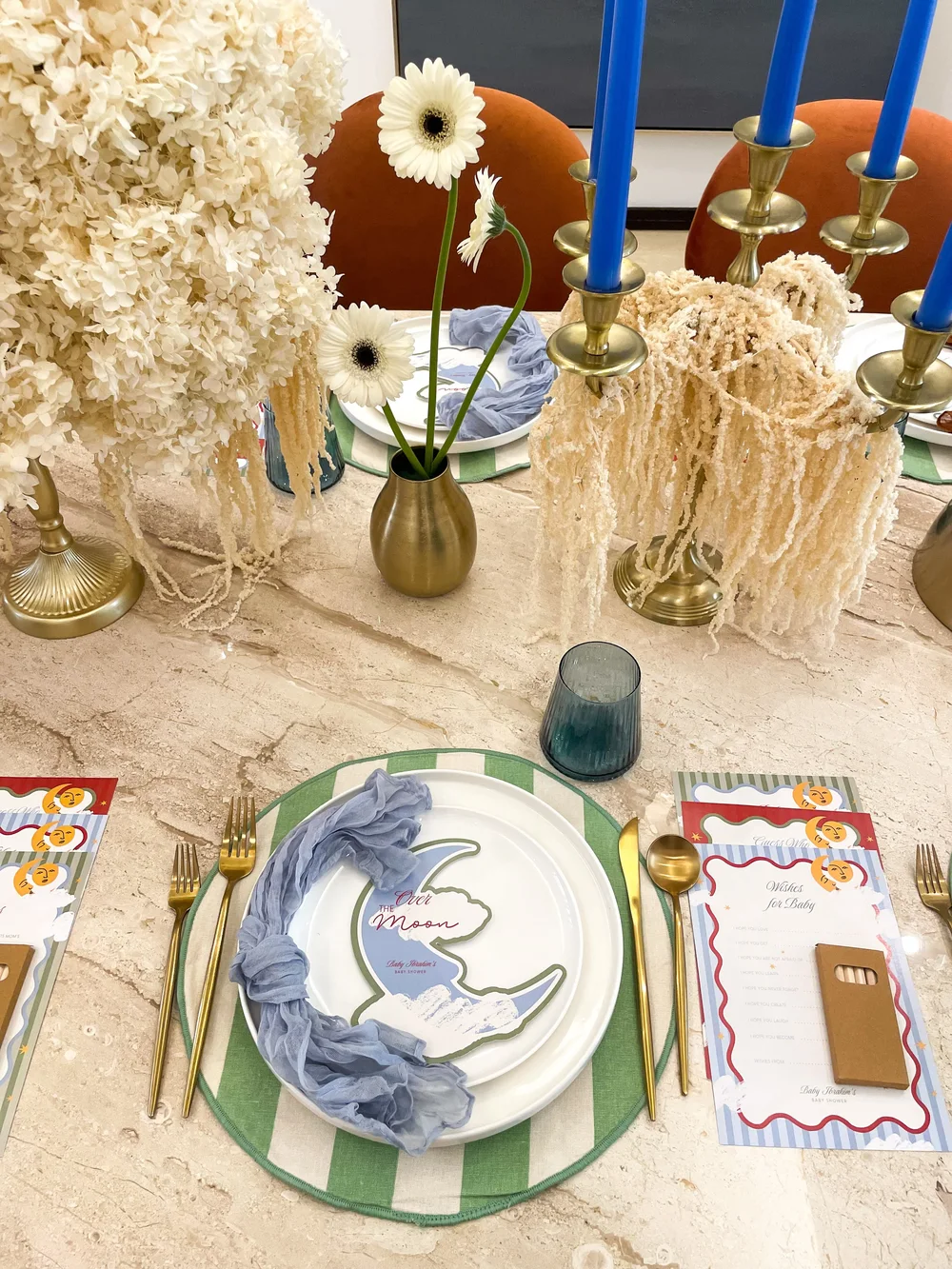 A baby shower place setting with a striped charger, gold cutlery and dried florals