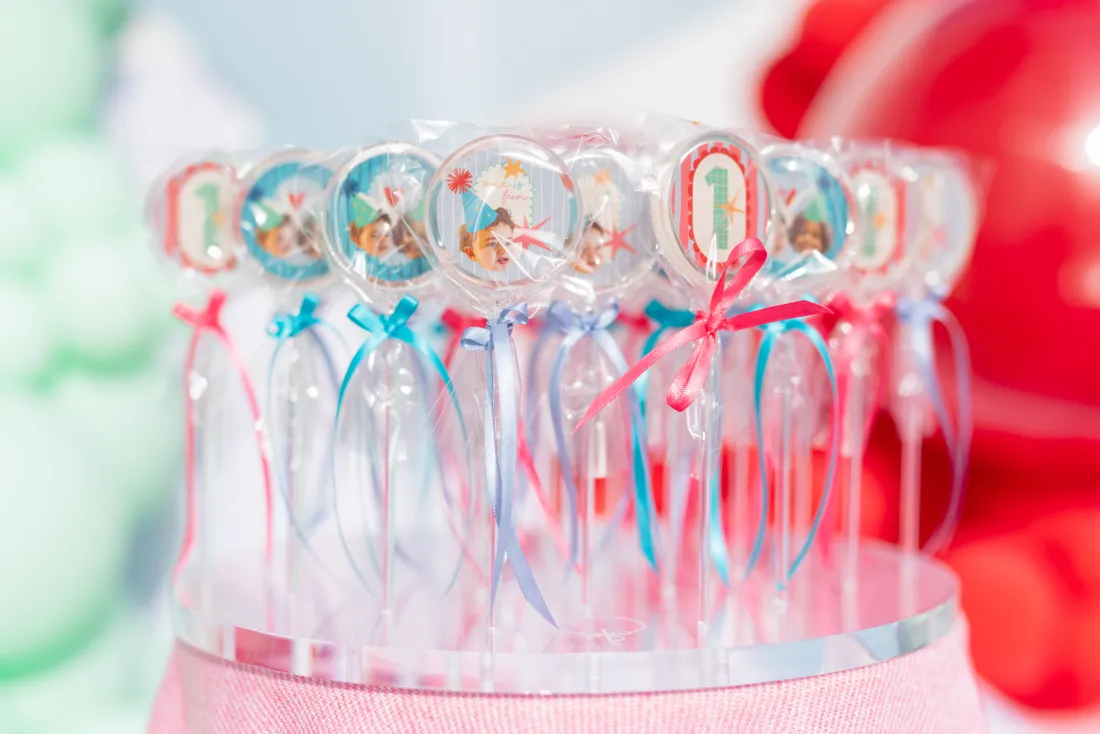 A row of personalised clear party favour wands with ribbons from a colourful first birthday setup