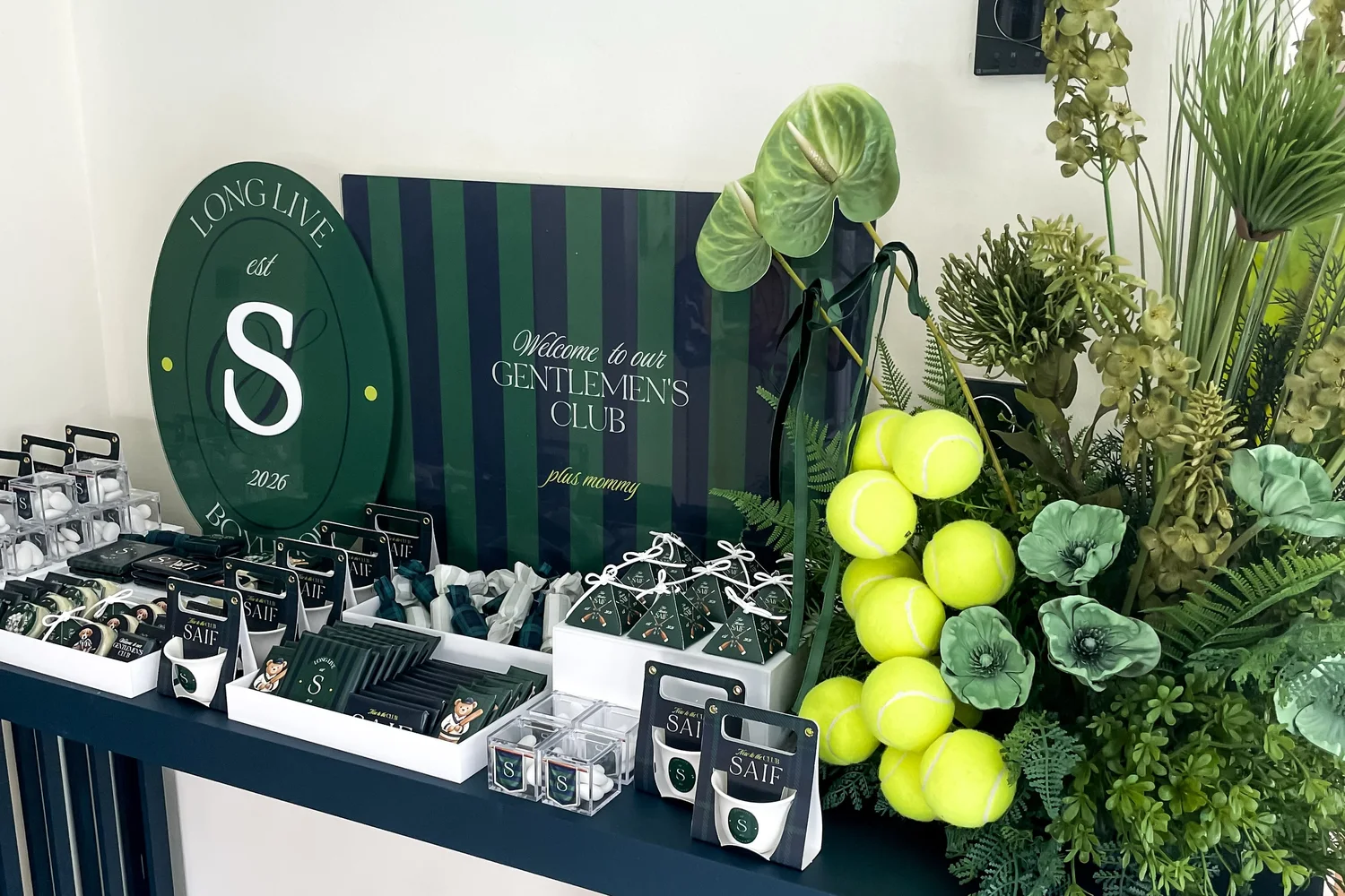 A gentlemen's club themed newborn welcoming with a dark green striped backdrop, tennis balls, greenery and personalised favours